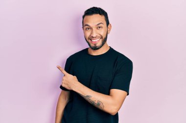 Sakallı, sıradan siyah tişörtlü, neşeli gülümseyen, eli ve parmağı yanda olan İspanyol bir adam. 