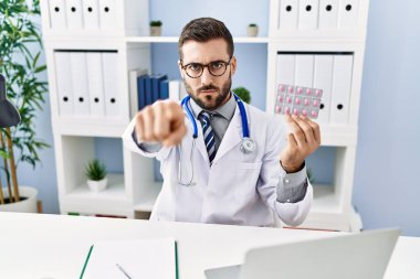 Yakışıklı, doktor üniforması giyen, elinde reçeteli haplar tutan, kamerayı işaret eden, kendine güvenen, ciddi görünen İspanyol bir adam. 