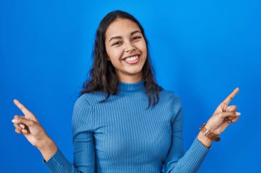 Mavi izole edilmiş arka planda duran genç Brezilyalı kadın farklı yönleri gösteren kendine güveniyle gülümsüyor. reklam için kopyalama alanı 