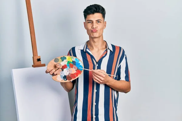 Joven Hombre Hispano Sosteniendo Paleta Pintor Pincel Pie Cerca Lienzo —  Fotos de Stock