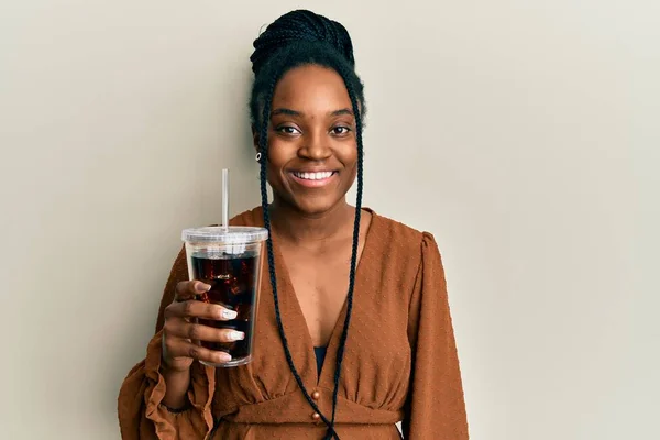African american woman with braided hair drinking glass of cola beverage looking positive and happy standing and smiling with a confident smile showing teeth