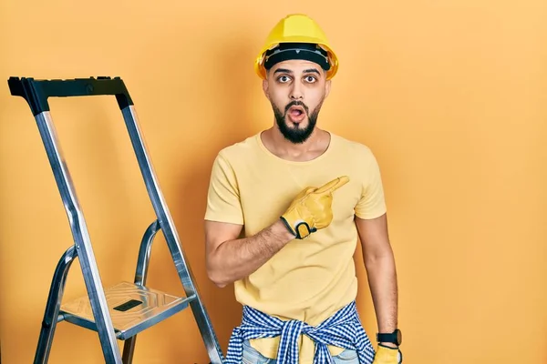 Handsome man with beard by construction stairs wearing hardhat surprised pointing with finger to the side, open mouth amazed expression.