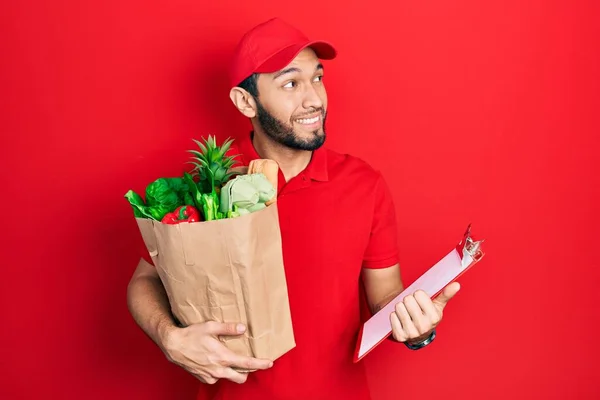 身穿信使制服的西班牙裔男子 身穿超市和剪贴板上的食品杂货 面带微笑地向旁边看去 目不转睛地想着 — 图库照片