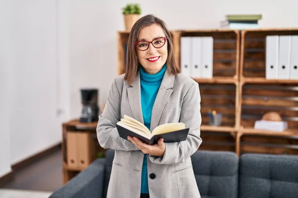 Middle age woman psychologist smiling confident reading book at psychology center
