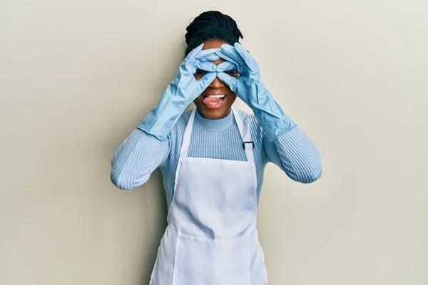 African american woman with braided hair wearing cleaner apron and gloves doing ok gesture like binoculars sticking tongue out, eyes looking through fingers. crazy expression.