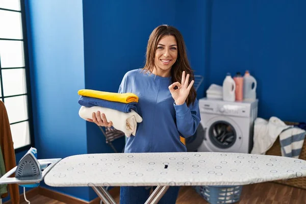 Young brunette woman holding folded laundry after ironing doing ok sign with fingers, smiling friendly gesturing excellent symbol