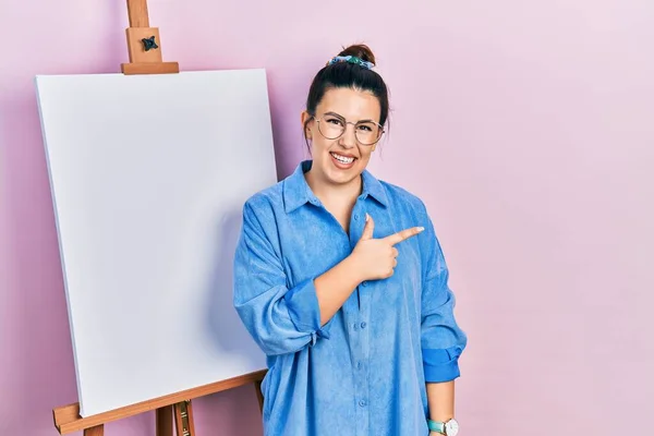 画家のイーゼルのそばに立つ若いヒスパニック系の女性は 幸せと自然な表情で側に手と指を指して笑顔で陽気に立っています — ストック写真