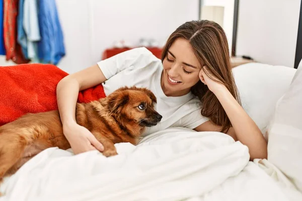 Young hispanic woman hugging dog lying on bed at bedroom