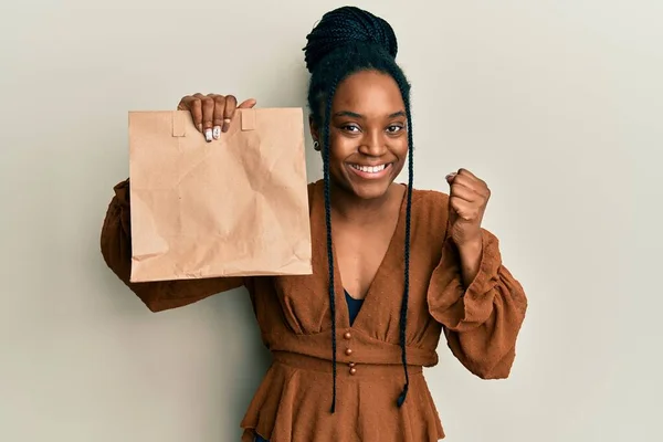 African american woman with braided hair holding take away paper bag screaming proud, celebrating victory and success very excited with raised arm