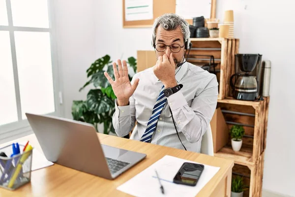 Middle age hispanic business man working at the office wearing operator headset smelling something stinky and disgusting, intolerable smell, holding breath with fingers on nose. bad smell