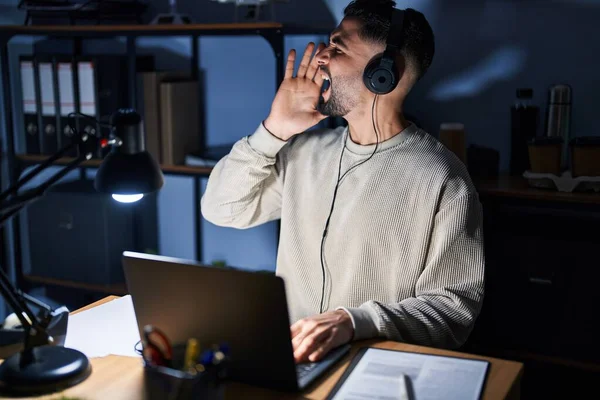 Young handsome man working using computer laptop at night shouting and screaming loud to side with hand on mouth. communication concept.