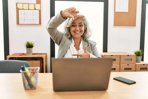 Middle age businesswoman sitting on desk working using laptop at office smiling making frame with hands and fingers with happy face. creativity and photography concept.