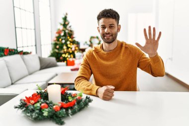 Arap genç adam Noel ağacının yanında oturuyor ve beş numarayı gösterip işaret ederken kendinden emin ve mutlu bir şekilde gülümsüyor.. 