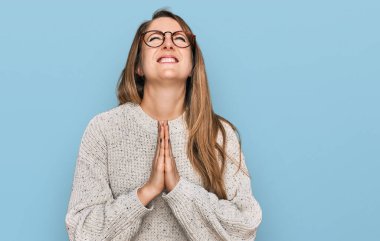 Sıradan süveter ve gözlük takan genç sarışın kadın elleri ile yalvarıyor ve dua ediyor. Yüzünde umut ifadesi var. Çok duygusal ve endişeli. Yalvarıyor. 