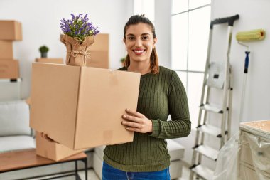 Genç İspanyol bir kadın, elinde karton bir kutuyla hareketli, pozitif ve mutlu görünüyor ve kendine güvenen bir gülümsemeyle dişlerini gösteriyor. 