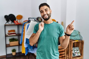 Perakende mağazasında sakallı yakışıklı bir adam. Yüzünde kocaman bir gülümsemeyle kameraya el ile bakıyor.. 