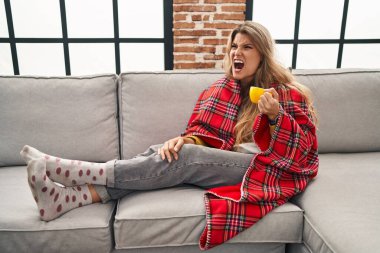 Kanepede oturan genç bir kadın kızgın ve kızgın bir şekilde bağırarak kahveyi yudumluyor öfkeyle bağırıyor. öfke ve agresif konsept. 