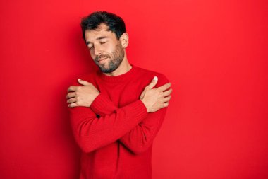 Sakallı, normal kırmızı süveter giyen, kendine sarılan mutlu ve pozitif, kendine güvenen yakışıklı bir adam. Sevgi ve özgüven. 