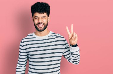 Sakallı, sıradan çizgili süveter giyen, kameraya gülümseyen ve parmaklarıyla zafer işareti yapan genç bir Arap. İki numara.. 