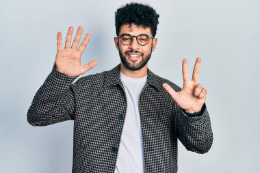 Sakallı, gözlüklü genç bir Arap sekiz numaralı parmaklarıyla gülümseyerek mutlu ve kendinden emin bir şekilde işaret ediyor.. 