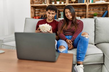 Laptopta film izleyen genç latin çift evdeki kanepede oturuyor..