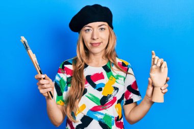 Kafkasyalı genç bir kadın sanatçı elinde küçük ahşap bir manikin ve yüzünde ciddi bir ifadeyle rahatlamış boya fırçaları tutuyor. Basit ve doğal bir şekilde kameraya bakıyorsun.. 