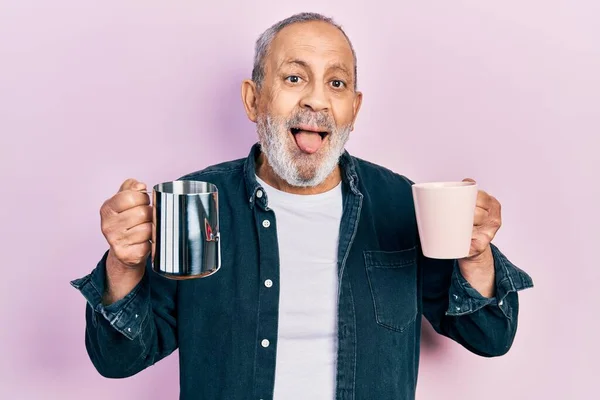 Handsome senior man with beard holding coffee and milk sticking tongue out happy with funny expression.