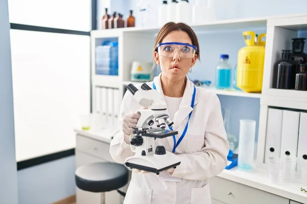 Young brunette woman working at scientist laboratory with microscope making fish face with mouth and squinting eyes, crazy and comical.