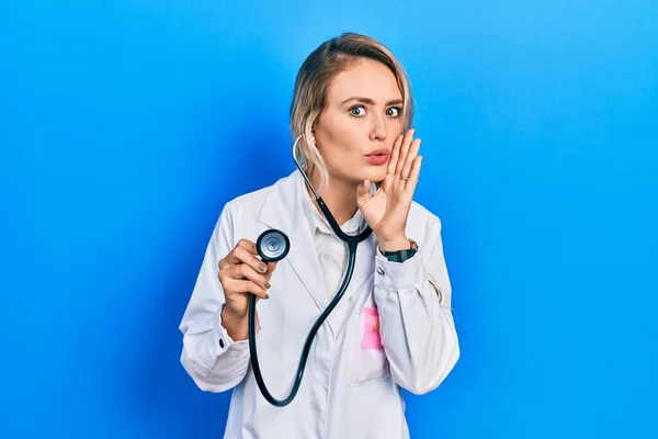 Beautiful young blonde doctor woman holding stethoscope hand on mouth telling secret rumor, whispering malicious talk conversation