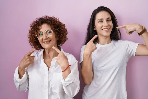 ピンクの背景にカジュアルな白いTシャツを着たヒスパニック系の母親と娘は 明るい表情と指の歯と口で指して笑っています 歯の健康の概念 — ストック写真
