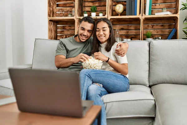 Young hispanic couple watching film and eating popcorn sitting on the sofa at home.