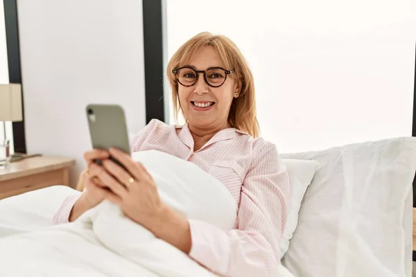 Middle age blonde woman using smartphone lying on the bed at home.