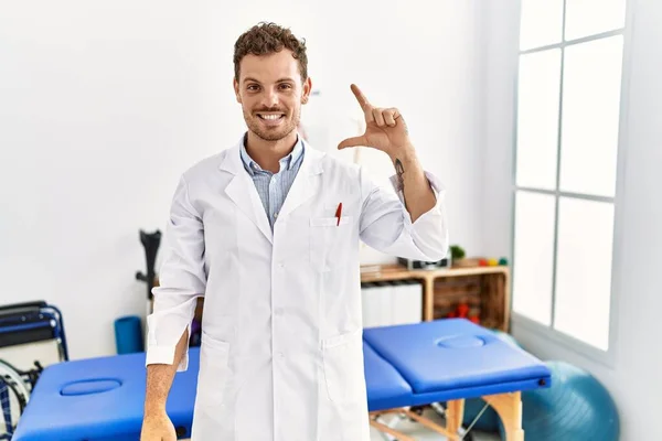 Handsome young man working at pain recovery clinic smiling and confident gesturing with hand doing small size sign with fingers looking and the camera. measure concept.