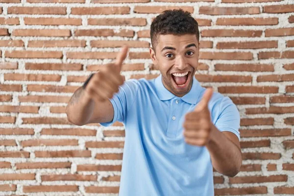 手で積極的なジェスチャーを行うことを承認レンガの壁の上に立ってブラジルの若い男 親指アップ笑顔と成功のために幸せ 勝者のジェスチャー — ストック写真
