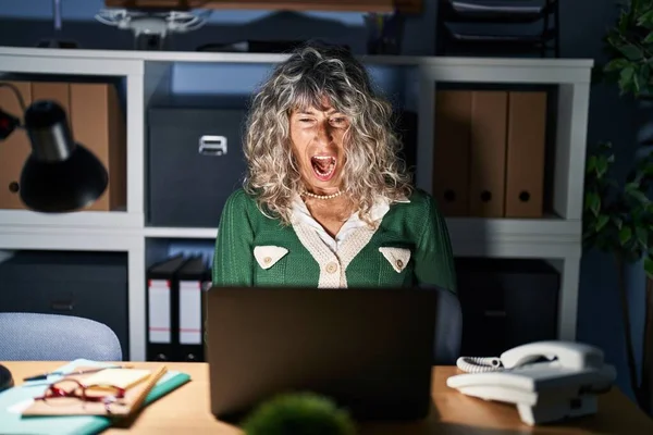 Middle age woman working at night using computer laptop angry and mad screaming frustrated and furious, shouting with anger. rage and aggressive concept.