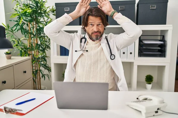 Handsome middle age doctor man working at the clinic doing bunny ears gesture with hands palms looking cynical and skeptical. easter rabbit concept.