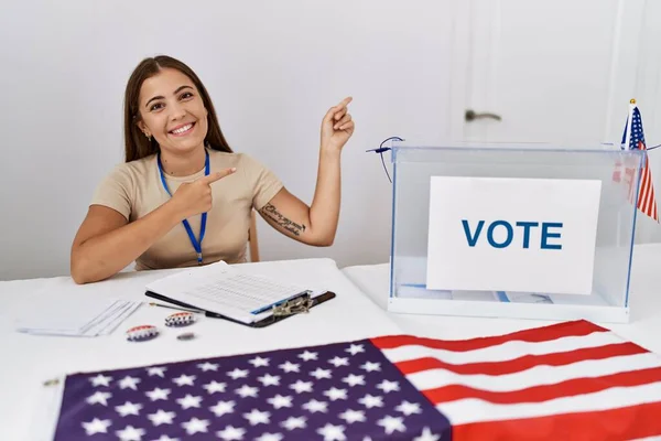 在政治选举中 年轻的黑发女子微笑着坐在选票旁 看着摄像机用双手和手指指向旁边 — 图库照片