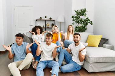Group of people sitting on the sofa and floor at home clueless and confused expression with arms and hands raised. doubt concept. 