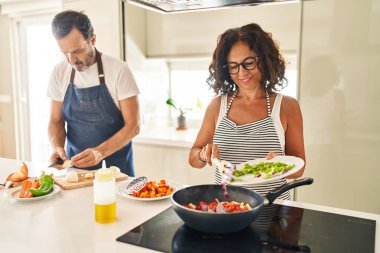 Orta yaşlı İspanyol çift, gülümsüyor. Mutfakta tavaya yiyecek koyuyorlar.