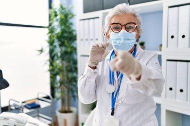 Gri saçlı, doktor üniforması giyen, elinde şırıngayla kameraya ve sana bakan yaşlı bir kadın. Kendine güvenen, ciddi bir hareket. 