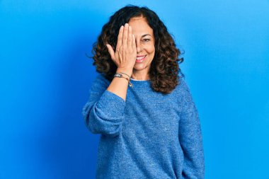 Orta yaşlı İspanyol kadın gündelik kıyafetler giyiyor. Bir gözünü eliyle kapatıyor. Yüzünde kendine güvenen bir gülümseme ve sürpriz bir duygu var.. 