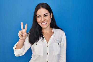 Mavi arka planda dikilen genç İspanyol kadın iki numaralı parmaklarını gösterip işaret ederken kendinden emin ve mutlu gülümsüyor.. 