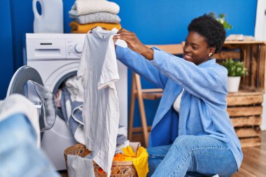 African american woman holding t shirt using washing machine at laundry room