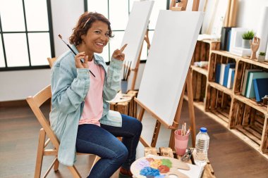 Orta yaşlı İspanyol kadın resim stüdyosunda tuvale resim yapıyor. Gülümsüyor, mutlu bir şekilde elini ve parmağını yana doğru gösteriyor. 
