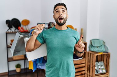 Satış mağazasında sakallı yakışıklı bir adam şaşırmış ve şaşırmış bir şekilde yukarı bakıyor ve parmaklarıyla işaret ediyor ve kollarını kaldırıyordu.. 