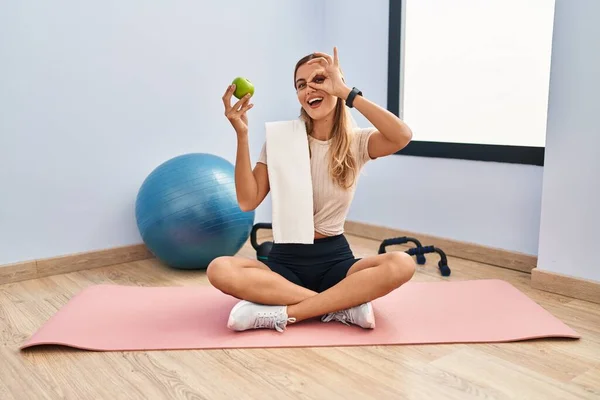 Young blonde woman wearing sportswear and towel holding healthy apple doing ok gesture with hand smiling, eye looking through fingers with happy face.
