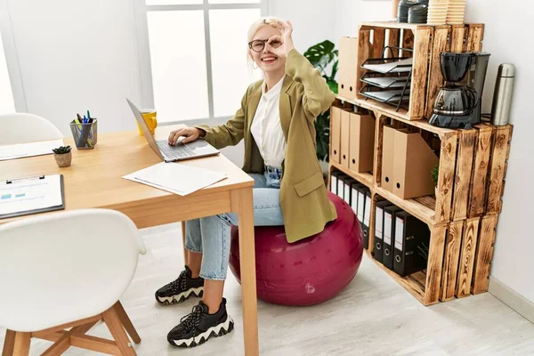 Beautiful caucasian business woman working at the office sitting on pilates ball smiling happy doing ok sign with hand on eye looking through fingers 