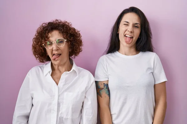 ヒスパニックの母親と娘はピンクの背景にカジュアルな白いTシャツを着て 面白い表情で幸せ舌を突き出しています 感情の概念 — ストック写真