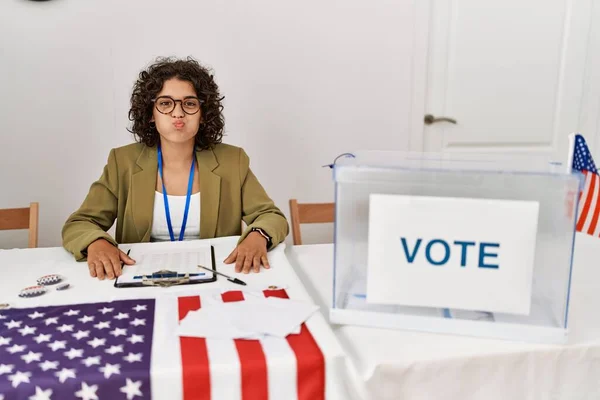 Young hispanic woman at political election sitting by ballot puffing cheeks with funny face. mouth inflated with air, crazy expression.