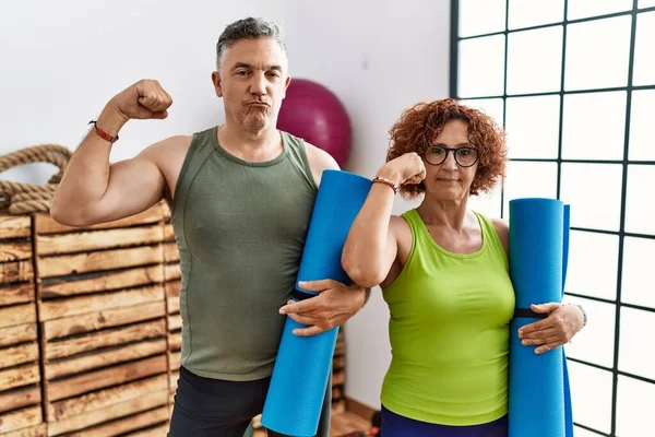 Middle age couple holding yoga mat strong person showing arm muscle, confident and proud of power